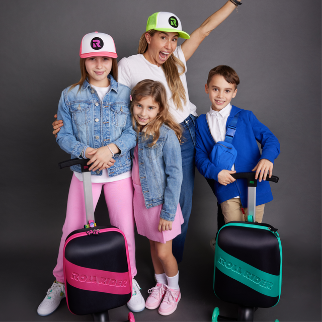 Jenny Fleiss smiling big standing with her three kids and a pink and a green Roll Rider scooter suitcase in a gray room.