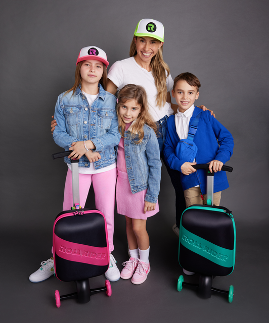 Jenny Fleiss standing and smiling with her three kids and a pink and a green Roll Rider scooter suitcase in a gray room.