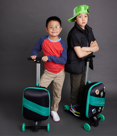 Two young boys posing casually with Roll Rider green and black scooter suitcases in a gray room. 