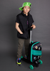 Young boy wearing a green cap backwards posing with a green Roll Rider scooter suitcase with stickers, in a gray room.