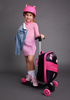 Smiling young girl wearing pink clothes and a pink helmet standing on a pink Roll Rider scooter suitcase in a gray room.